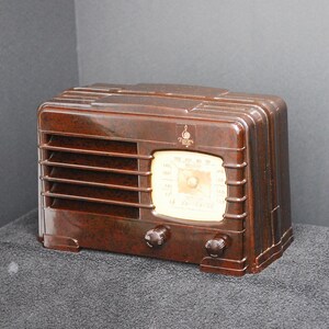 May include: A vintage brown radio with a glossy finish. The radio features a cream-colored dial with black markings and two dark knobs. The front has horizontal slats and a small logo. A classic piece of home decor.