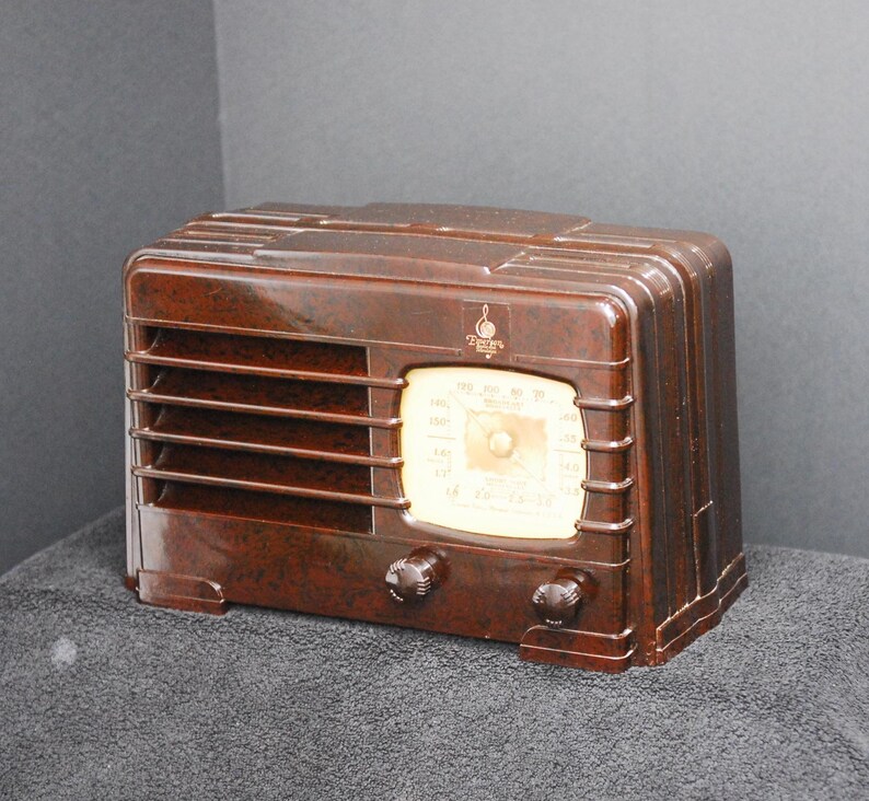 May include: A vintage brown radio with a glossy finish. The radio features a cream-colored dial with black markings and two dark knobs. The front has horizontal slats and a small logo. A classic piece of home decor.