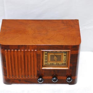 May include: A vintage wooden radio with a warm, brown finish. The radio has a rectangular design with rounded edges and a speaker grill on the left. It features a rectangular display with a dial and three black knobs at the bottom.