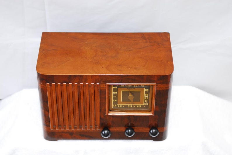 May include: A vintage wooden radio with a warm, brown finish. The radio has a rectangular design with rounded edges and a speaker grill on the left. It features a rectangular display with a dial and three black knobs at the bottom.
