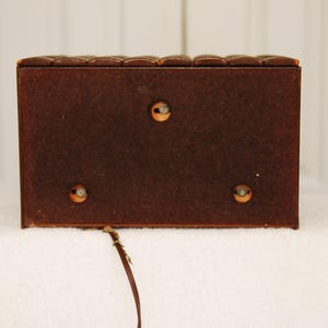 May include: A brown wooden box with a textured top and three visible screws on the back. The box is sitting on a white surface.