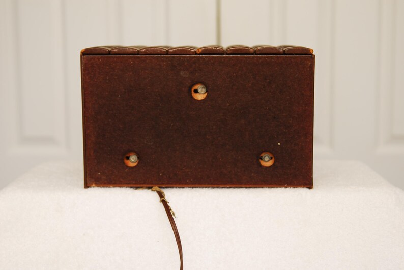 May include: A brown wooden box with a textured top and three visible screws on the back. The box is sitting on a white surface.