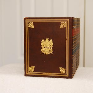 May include: A set of brown leather-bound books with gold accents and a crest on the front cover.