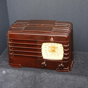 May include: A vintage brown radio with a cream-colored dial and two knobs. The radio has a rectangular shape with horizontal vents and a decorative top. The dial displays numbers and a needle, and the radio is set on a dark surface.
