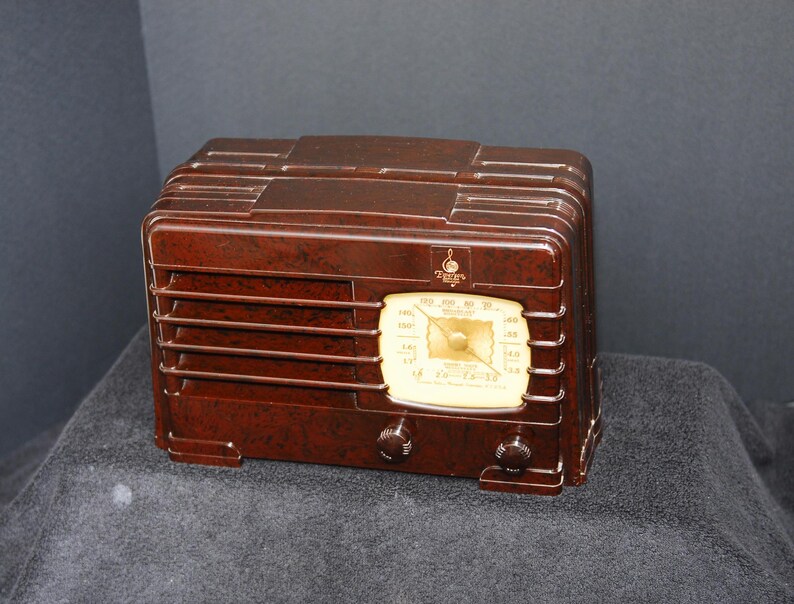 May include: A vintage brown radio with a cream-colored dial and two knobs. The radio has a rectangular shape with horizontal vents and a decorative top. The dial displays numbers and a needle, and the radio is set on a dark surface.