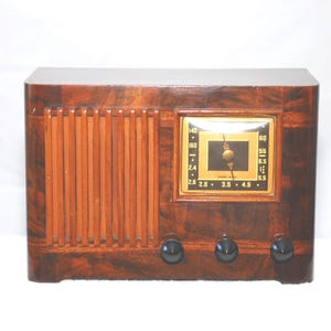 May include: A vintage wooden radio with a dark brown finish. The radio has a rectangular design with vertical wooden slats on the left and a square clock face in the centre. Three black knobs are at the bottom.