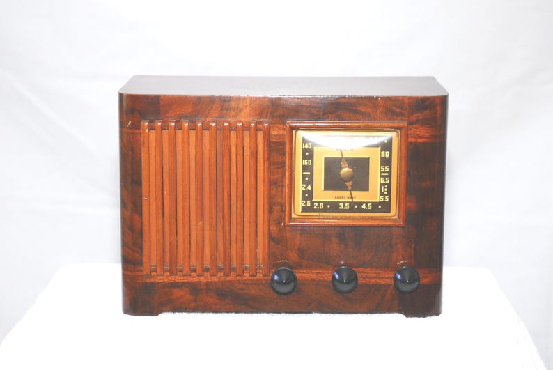 May include: A vintage wooden radio with a dark brown finish. The radio has a rectangular design with vertical wooden slats on the left and a square clock face in the centre. Three black knobs are at the bottom.
