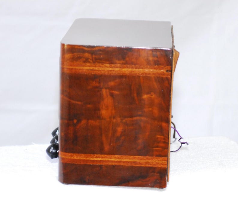 May include: A vintage wooden radio with a glossy, dark brown finish. The radio has a rectangular shape with rounded edges and a contrasting lighter wood band. Black knobs and wires are visible on the side.