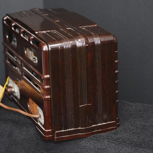 May include: A vintage brown radio with a glossy finish. The radio has a rectangular shape with vertical and horizontal design elements. A yellow and brown cord extends from the side. The radio is likely from the mid-20th century.