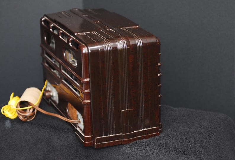 May include: A vintage brown radio with a glossy finish. The radio has a rectangular shape with vertical and horizontal design elements. A yellow and brown cord extends from the side. The radio is likely from the mid-20th century.