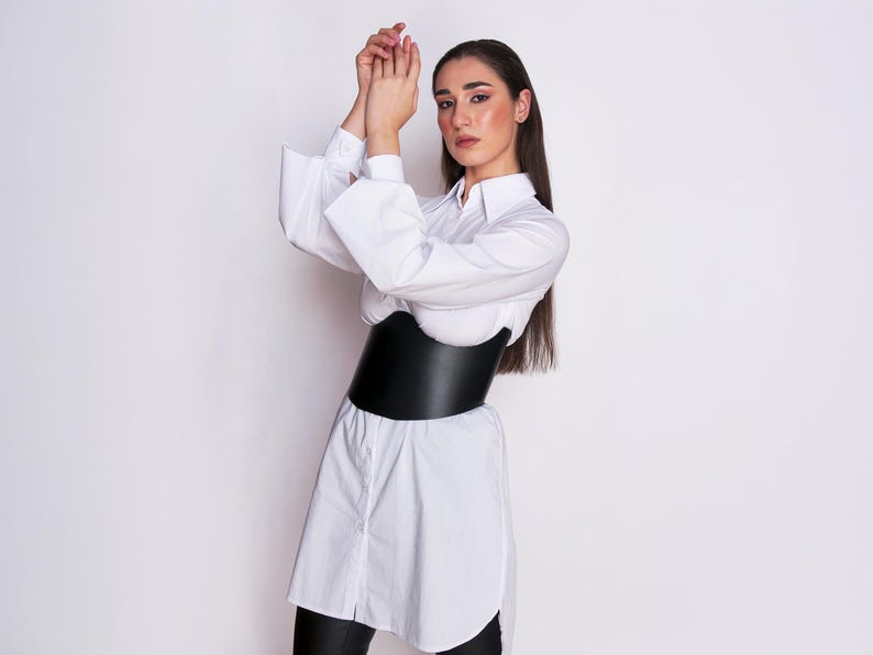 A model wearing a structured black leather underbust corset belt over an oversized white button-down shirt shirt, displaying a side view of the smooth leather and dramatic silhouette framing.