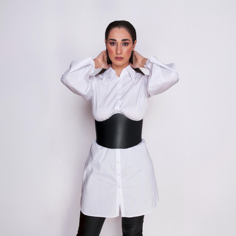 Front studio portrait of a model wearing a wide black leather underbust corset belt cinched over a white tailored shirt dress.