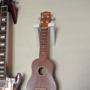 May include: A brown and gold electric guitar with a strap hanging from it is mounted on a wall next to a smaller brown ukulele.