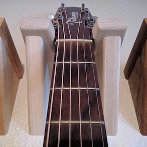 May include: A close-up of a guitar headstock with six strings, resting in a wooden guitar stand. The stand is made of light wood and has a triangular shape. The guitar is a light brown color with a dark brown fretboard.