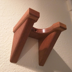 May include: A wooden wall mount for hanging a guitar. The mount is made of dark brown wood and has a triangular shape. It has a round hole in the center for the guitar's neck.