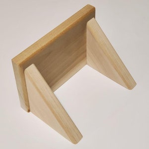 May include: A small, unfinished wooden stand with a rectangular top and two triangular supports. The natural wood has a light, warm tone, and the stand is designed to hold an item upright.