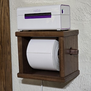May include: A white Rollo thermal label printer with purple accents sits atop a dark brown wooden box holder. Inside the box, a large roll of white adhesive labels is mounted on a horizontal wooden dowel, allowing the labels to feed through the unit.