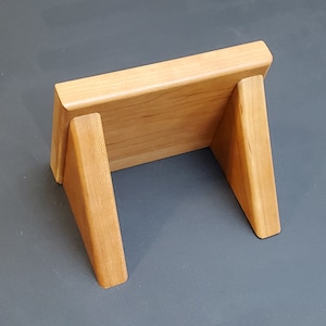 May include: A wooden display stand with a rectangular top and two angled sides. The stand is made of light brown wood and has a natural finish.