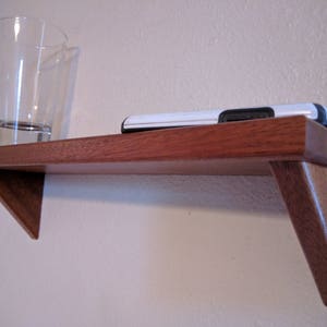 May include: A wooden shelf with a triangular design, mounted on a wall. Two clear glass cups are on the shelf, and a black and white cell phone is resting on the shelf.