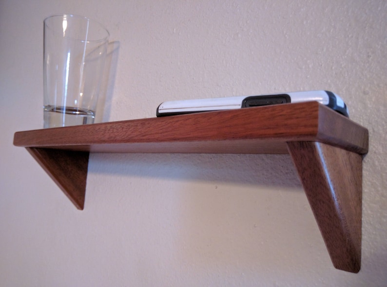 May include: A wooden shelf with a triangular design, mounted on a wall. Two clear glass cups are on the shelf, and a black and white cell phone is resting on the shelf.