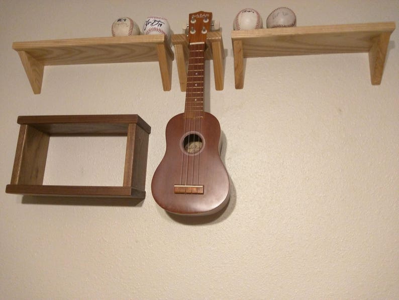 May include: A wooden shelf unit with three shelves, a small shelf on the left, a larger shelf on the right, and a smaller shelf in the middle. A ukulele is hanging from the middle shelf. There are three baseballs on the top shelf.