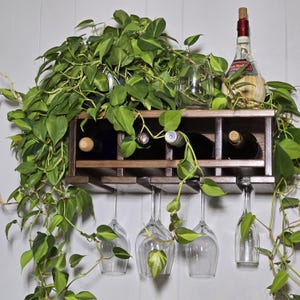 May include: A wooden wine rack with four bottles and four wine glasses. A bottle of wine with a cork is on top of the rack. Green leafy vines are draped over the rack and glasses.