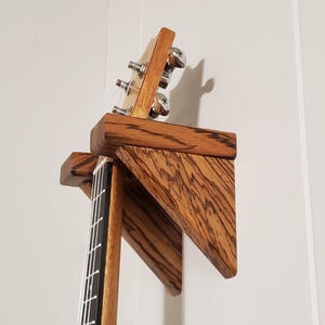 May include: A wooden wall mount guitar stand with a dark brown wood grain finish. The stand is designed to hold a guitar by the neck, with a triangular shape to support the instrument.