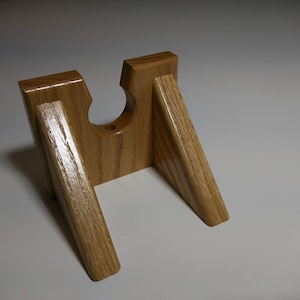 May include: A wooden stand with a cutout for a bottle or other object. The stand is made of light brown wood and has a simple design.