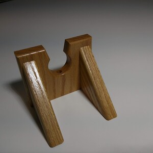 May include: A wooden stand with a cutout for holding a small object, such as a phone or a tablet. The stand is made of light brown wood and has a simple, modern design.
