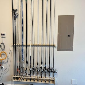 May include: A collection of fishing rods of varying lengths and designs, mounted on a wooden rack against a light-colored wall. The rods are black, white, and blue, with green tips. The rack is mounted near an electrical panel and other equipment.