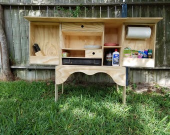 Puede incluir: Un armario de madera con estantes y cajones, ideal para acampar. Tiene una encimera y un porta rollo de papel. El armario está abierto, mostrando el interior.