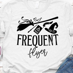 以下が含まれることがあります： 白いTシャツに、ほうき、メガネ、羽、魔女の帽子、そして「FREQUENT flyer」の文字が描かれた黒いグラフィックデザイン。シャツはブルージーンズと白いスニーカーと組み合わせています。デジタルダウンロード。