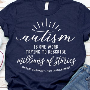 May include: A navy blue t-shirt with white text that reads "autism is one word trying to describe millions of stories offer support, not judgement". The text is in a stylized font with a sunburst design above the word "autism".