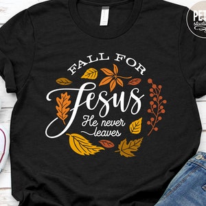 Puede incluir: Una camiseta negra con un diseño gráfico blanco que dice "Fall for Jesus He never leaves". El diseño gráfico está rodeado de hojas de otoño en tonos de naranja, amarillo y rojo.