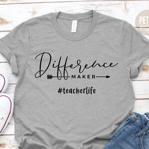 Peut inclure: Un t-shirt gris avec le texte "Difference Maker" et une flèche pointant vers la droite. En dessous du texte se trouve le hashtag "#teacherlife".