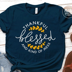 May include: A navy blue t-shirt with a white circle design that says "Thankful Blessed and Kind of Mess" with a gold leaf design around the words.