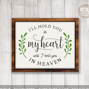 Puede incluir: Un letrero blanco con marco de madera marrón, con el texto "I'll hold you in my heart until I hold you in heaven." El texto está en fuente cursiva negra con brotes de hojas verdes a cada lado.