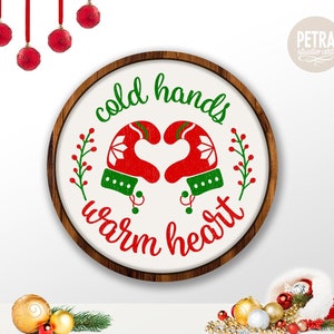 May include: A round wooden sign with the text "cold hands warm heart" in red and green lettering. The sign features a pair of red mittens with green trim forming a heart shape.