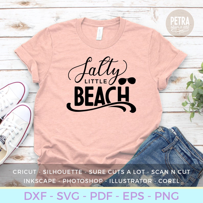 May include: A pink t-shirt with the text "Salty Little Beach" in black with sunglasses graphic. The text is in a cursive font. The shirt is on a wooden surface with a pair of white sneakers and blue jeans.