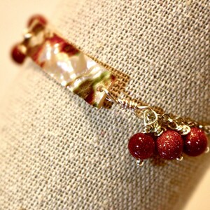 Peut inclure: Un bracelet en or avec un charme à motif blanc et vert et des perles rouges. Le bracelet est sur un fond en tissu beige.