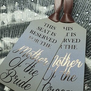 May include: Two grey place cards with gold lettering. The text reads "This seat is reserved for the Mother of the Bride" and "This seat is reserved for the Mother of the Groom".