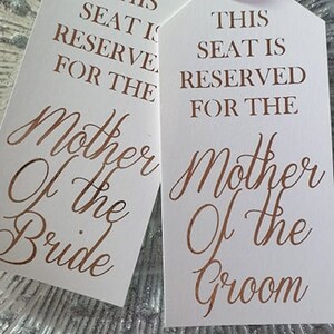 May include: Two white wedding table place cards with pink ribbon ties. The text on the cards reads "This seat is reserved for the Mother of the Bride" and "This seat is reserved for the Mother of the Groom".