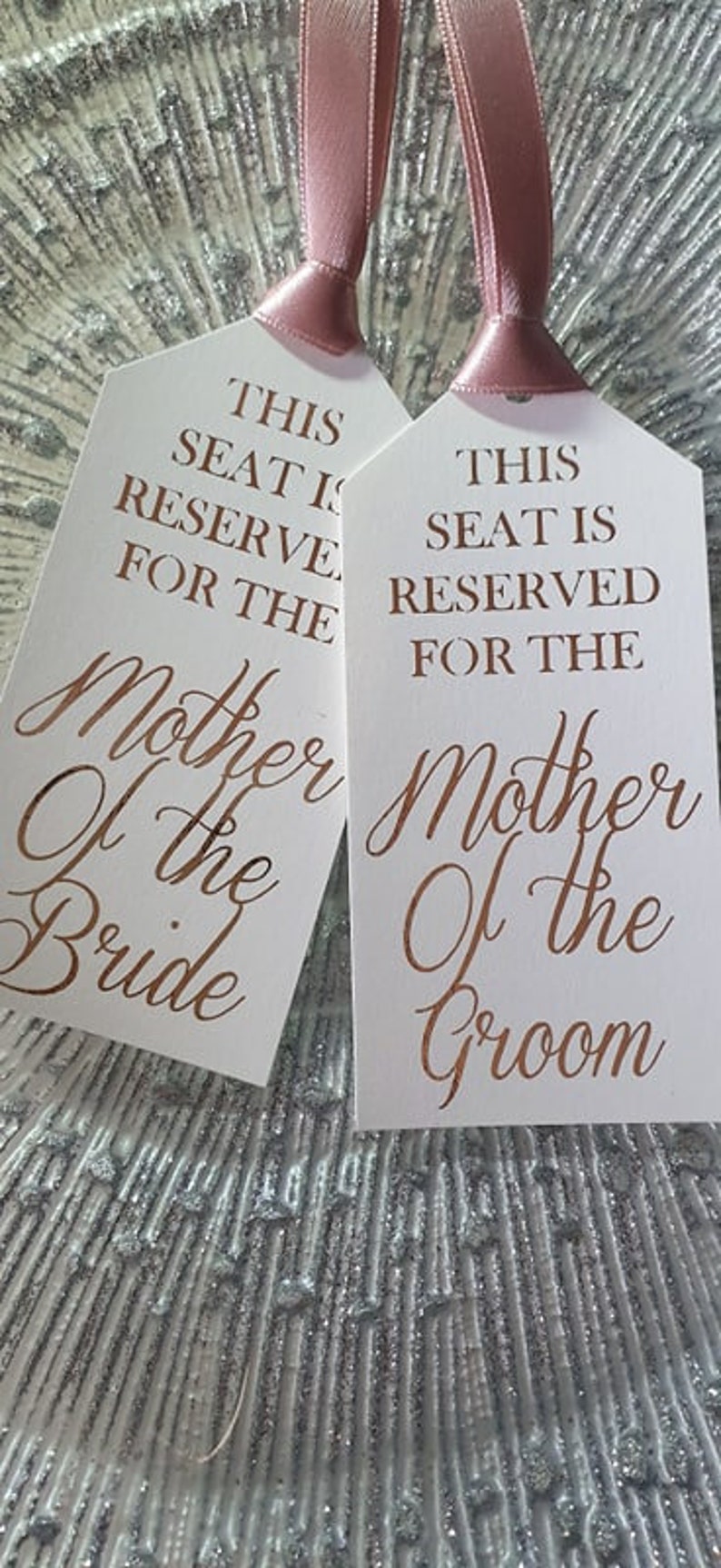 May include: Two white wedding table place cards with pink ribbon ties. The text on the cards reads "This seat is reserved for the Mother of the Bride" and "This seat is reserved for the Mother of the Groom".