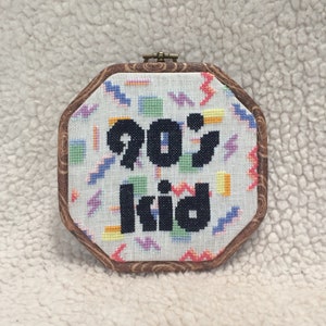 May include: A framed cross-stitch with a white background and a colorful geometric pattern. The text "90's kid" is stitched in black.