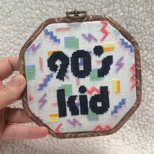 May include: A framed cross-stitch with a white background and colorful geometric patterns. The text "90's kid" is stitched in black.