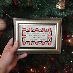 May include: A framed cross-stitch with a red, green, and yellow floral border. The text reads "And Mom and Dad can hardly wait for school to start again."