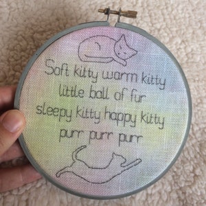 May include: A circular embroidery hoop with a rainbow gradient background. The hoop features a black and white embroidered design of a cat with the text "Soft kitty warm kitty little ball of fur sleepy kitty happy kitty purr purr purr".