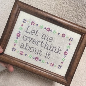 May include: A framed cross-stitch with a white background and a gray embroidered message that reads "Let me overthink about it". The embroidery is surrounded by a border of small colorful flowers.