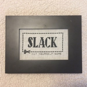 May include: A black framed cross-stitch with white fabric and black thread. The cross-stitch says "SLACK CUT YOURSELF SOME".