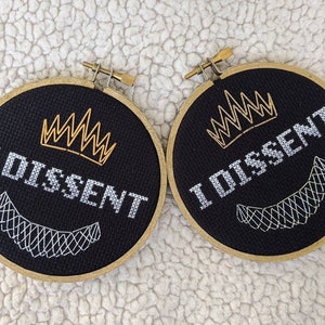 May include: Two black embroidery hoops with gold thread. The hoops feature a crown and the text "I Dissent".